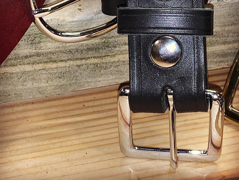 Peut inclure: Une ceinture en cuir noir avec une boucle argent&eacute;e et un bouton argent&eacute;. La ceinture est sur une surface en bois.