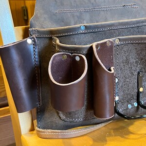 May include: A brown leather tool belt with multiple pockets and a metal hook. The belt has a strap and is made of a durable material.