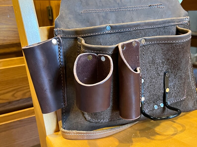 May include: A brown leather tool belt with multiple pockets and a metal hook. The belt has a strap and is made of a durable material.