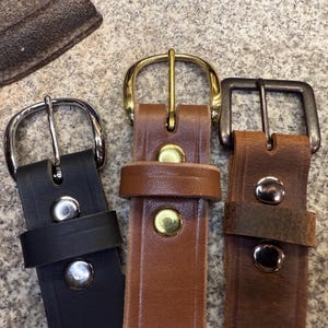 May include: Three leather belts with varying buckle designs. The belts are black, mid-brown, and dark brown. The buckles are silver, gold, and gunmetal. Each belt features metal rivets securing the leather.