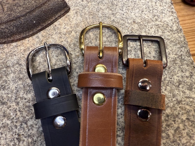 May include: Three leather belts with varying buckle designs. The belts are black, mid-brown, and dark brown. The buckles are silver, gold, and gunmetal. Each belt features metal rivets securing the leather.