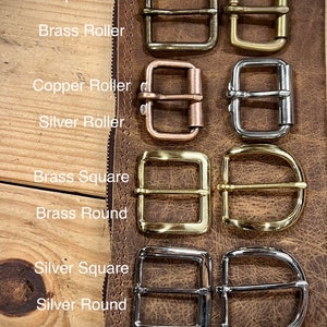 May include: An assortment of metal buckles in different styles and finishes, including antique brass, brass, copper, and silver. The buckles are square, round, and roller styles, presented on a brown leather surface. The image text reads "Antique Brass", "Brass Roller", "Copper Roller", "Silver Roller", "Brass Square", "Brass Round", "Silver Square", and "Silver Round".