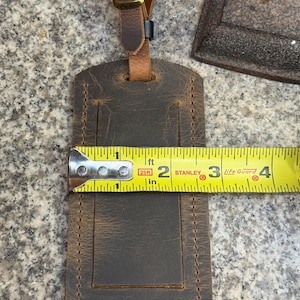 Op de afbeelding: Een bruine leren bagagelabel met een gestikte rand, ongeveer 10 cm lang. Het heeft een gesp en een riem voor bevestiging. Het label wordt getoond met een meetlint.