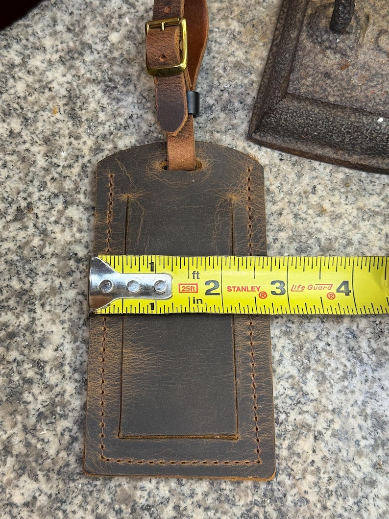 Op de afbeelding: Een bruine leren bagagelabel met een gestikte rand, ongeveer 10 cm lang. Het heeft een gesp en een riem voor bevestiging. Het label wordt getoond met een meetlint.