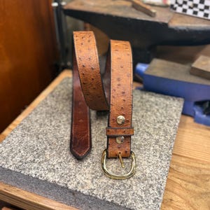 May include: A tan belt with a textured ostrich-like pattern, featuring a polished gold-tone metal buckle and two matching gold-tone rivets securing the strap near the buckle. The belt has a pointed tip and a smooth, darker brown interior lining.