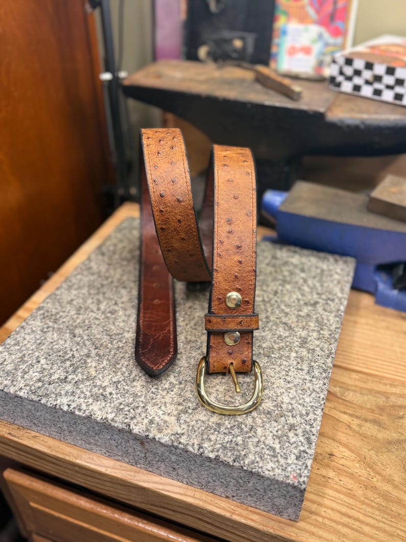 May include: A tan belt with a textured ostrich-like pattern, featuring a polished gold-tone metal buckle and two matching gold-tone rivets securing the strap near the buckle. The belt has a pointed tip and a smooth, darker brown interior lining.