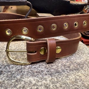 May include: A brown leather belt with a brass buckle and brass eyelets. The belt has a classic design and is likely handmade. The buckle is a simple, curved design. The belt is shown on a textured surface.