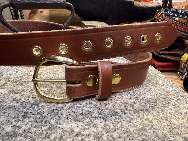 May include: A brown leather belt with a brass buckle and brass eyelets. The belt has a classic design and is likely handmade. The buckle is a simple, curved design. The belt is shown on a textured surface.