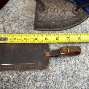 Op de afbeelding: Een bruine leren bagagelabel met een gespriem, ongeveer 10 cm breed. De label ligt naast een gele rolmaat en een antieke strijkijzer op een stenen oppervlak. De label heeft een rechthoekige vorm met een gleuf voor een identificatiekaart.
