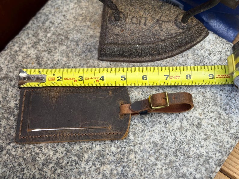 Op de afbeelding: Een bruine leren bagagelabel met een gespriem, ongeveer 10 cm breed. De label ligt naast een gele rolmaat en een antieke strijkijzer op een stenen oppervlak. De label heeft een rechthoekige vorm met een gleuf voor een identificatiekaart.