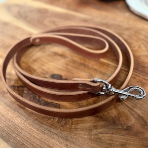 May include: A rolled-up brown leather dog leash with a silver-colored metal clasp. The leash is made of a single strip of leather and is lying on a wooden surface.