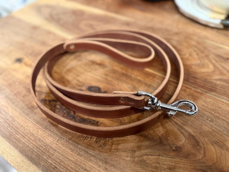 May include: A rolled-up brown leather dog leash with a silver-colored metal clasp. The leash is made of a single strip of leather and is lying on a wooden surface.