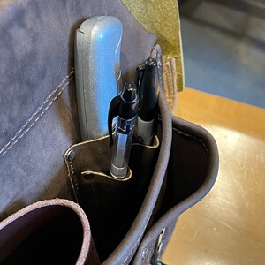 May include: A brown leather tool belt with a silver utility knife and two black pens in the pockets.