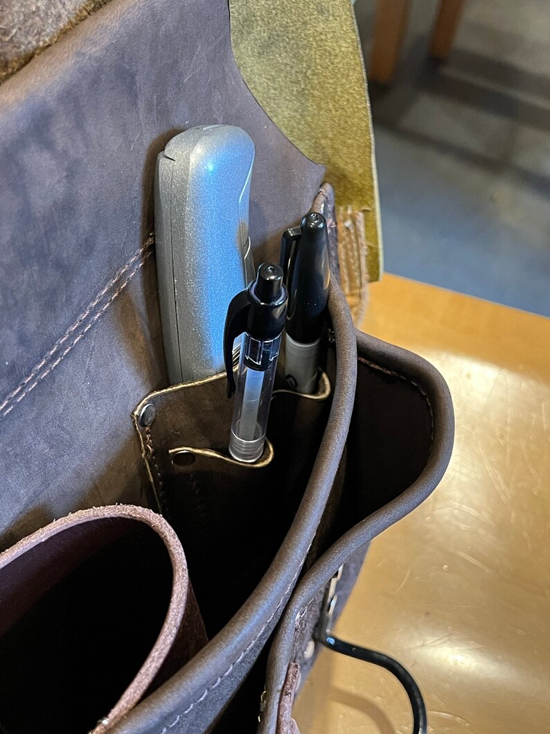 May include: A brown leather tool belt with a silver utility knife and two black pens in the pockets.