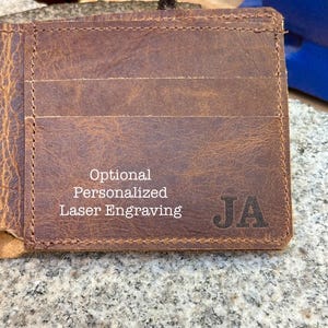 Może przedstawiać: Brązowy sk&oacute;rzany portfel z wieloma przegr&oacute;dkami na karty. Portfel ma inicjały "JA" wygrawerowane z przodu. Tekst na portfelu brzmi "Optional Personalized Laser Engraving".
