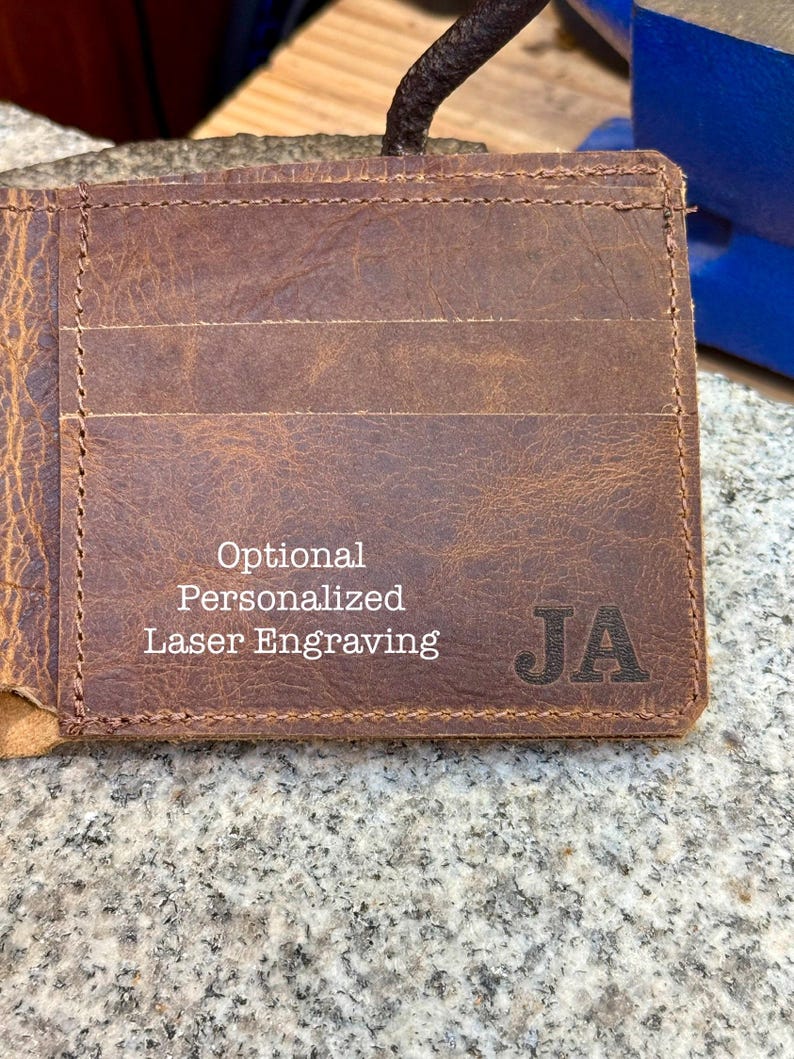 Może przedstawiać: Brązowy sk&oacute;rzany portfel z wieloma przegr&oacute;dkami na karty. Portfel ma inicjały "JA" wygrawerowane z przodu. Tekst na portfelu brzmi "Optional Personalized Laser Engraving".