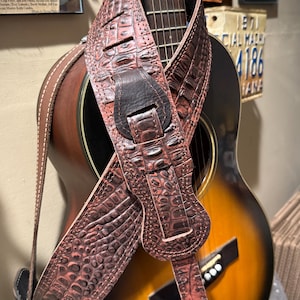 May include: Acoustic guitar with a brown and black leather guitar strap. The strap has a textured, crocodile-like pattern. The guitar has a sunburst finish and a black pickguard.