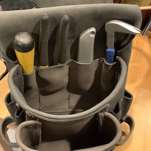 May include: A dark gray leather tool pouch featuring multiple pockets and loops. The pouch contains a yellow-handled screwdriver, two black-handled pliers, a silver utility knife, and a silver metal pry bar with a blue handle.