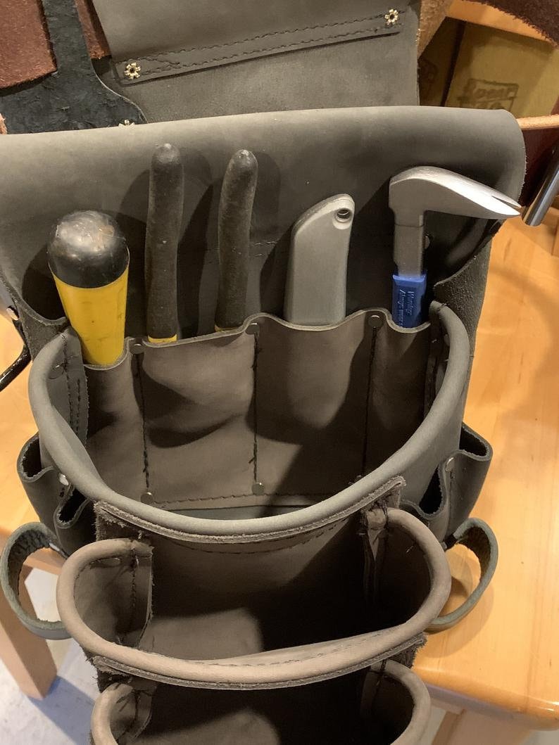 May include: A dark gray leather tool pouch featuring multiple pockets and loops. The pouch contains a yellow-handled screwdriver, two black-handled pliers, a silver utility knife, and a silver metal pry bar with a blue handle.