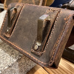 May include: A brown leather case with two metal clips and a stitched edge. The case is worn and has a distressed look.