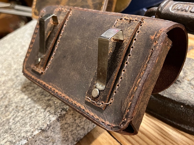 May include: A brown leather case with two metal clips and a stitched edge. The case is worn and has a distressed look.