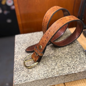 May include: A tan belt with a textured, dotted surface pattern, featuring a polished gold-tone metal buckle and a matching metal stud on the keeper loop. The belt has dark brown edges and a smooth, dark brown interior lining visible in the loops.