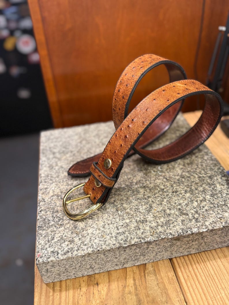 May include: A tan belt with a textured, dotted surface pattern, featuring a polished gold-tone metal buckle and a matching metal stud on the keeper loop. The belt has dark brown edges and a smooth, dark brown interior lining visible in the loops.