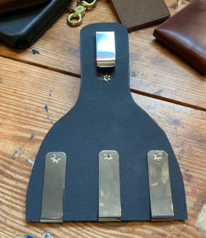 May include: A black leather wallet with three metal clips and a single metal clip on the top. The clips are secured with silver screws.