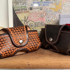 May include: Two leather cases with snap closures. One is a light brown with a woven pattern, and the other is dark brown. Both have white stitching and are displayed on a wooden surface. The background includes a framed poster with text.