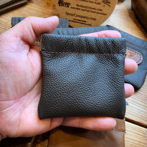 Peut inclure: Une petite pochette en cuir marron fonc&eacute; avec un d&eacute;tail de couture marron. La pochette est tenue dans une main.