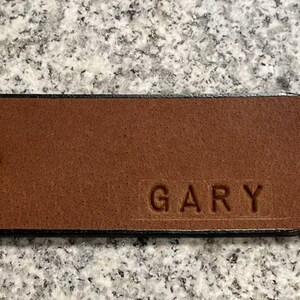 Pu&ograve; includere: Una cintura in pelle marrone con un bordo nero e un solo foro. La cintura &egrave; personalizzata con il nome "GARY" inciso nella pelle.