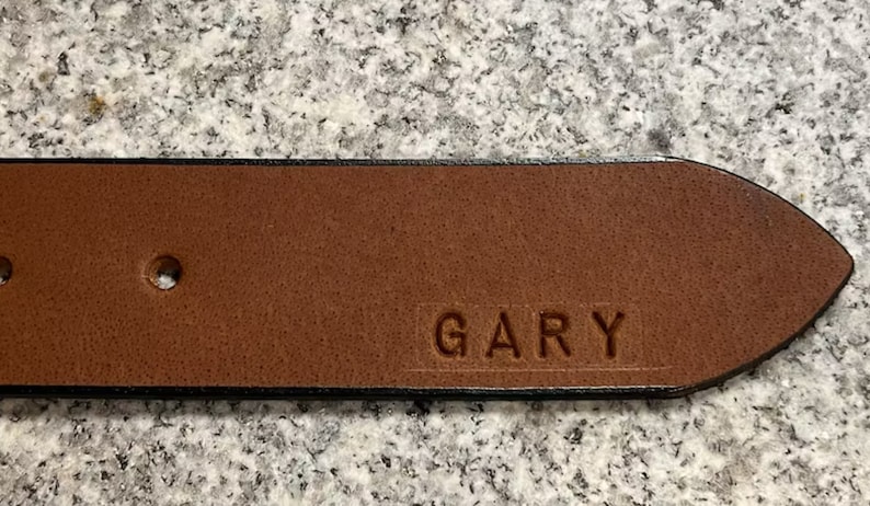 Pu&ograve; includere: Una cintura in pelle marrone con un bordo nero e un solo foro. La cintura &egrave; personalizzata con il nome "GARY" inciso nella pelle.