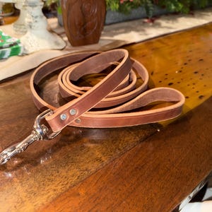 May include: A coiled brown leather dog leash with a silver-colored clasp. The leash is made of a single strip of leather and is secured with rivets. The leash is lying on a wooden surface.