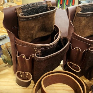 May include: Two brown leather tool belts with multiple pockets and compartments. The belts have a worn, vintage look and are made of high-quality leather.