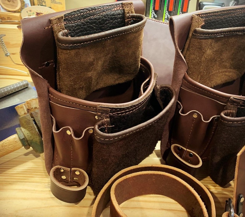 May include: Two brown leather tool belts with multiple pockets and compartments. The belts have a worn, vintage look and are made of high-quality leather.