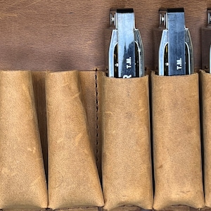 Puede incluir: Primer plano de tres arm&oacute;nicas en una funda de cuero marr&oacute;n. Cada arm&oacute;nica tiene un cuerpo plateado y una parte superior de pl&aacute;stico negro con el texto "T.M." impreso en ella.
