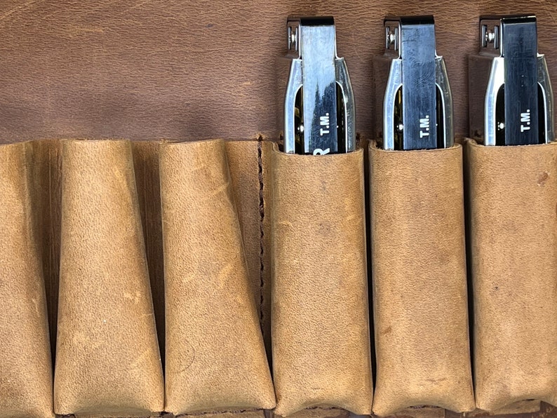 Puede incluir: Primer plano de tres arm&oacute;nicas en una funda de cuero marr&oacute;n. Cada arm&oacute;nica tiene un cuerpo plateado y una parte superior de pl&aacute;stico negro con el texto "T.M." impreso en ella.