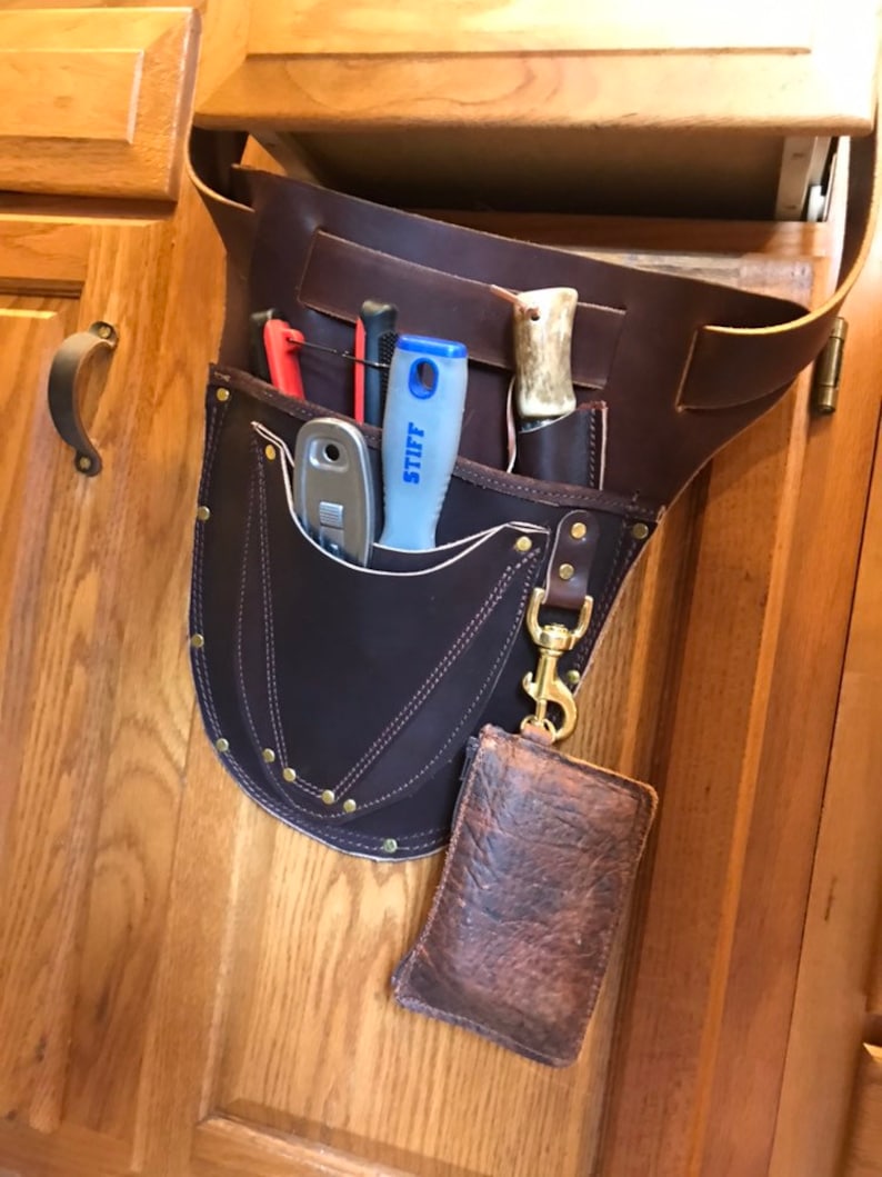 May include: A brown leather tool belt with multiple pockets and a small pouch hanging from it. The belt is hanging on a wooden cabinet door. The pockets contain various tools, including a knife, a screwdriver, and a utility knife. The pouch is made of brown leather and has a flap closure.