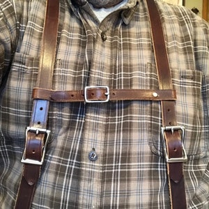 May include: A pair of dark brown leather suspenders featuring two vertical straps connected by a horizontal chest strap. The suspenders include silver-toned metal buckles for adjustment and secure fastening, worn over a grey and white plaid button-down shirt.