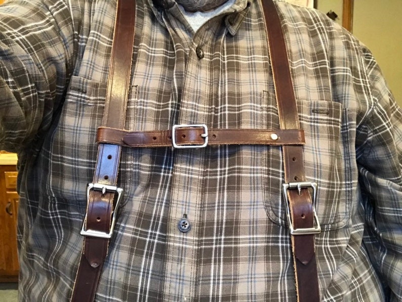 May include: A pair of dark brown leather suspenders featuring two vertical straps connected by a horizontal chest strap. The suspenders include silver-toned metal buckles for adjustment and secure fastening, worn over a grey and white plaid button-down shirt.