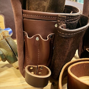 May include: A brown leather tool belt with multiple pockets and a suede flap. The belt has a metal buckle and is designed for carrying tools.