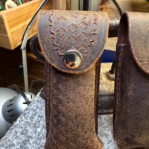K&ouml;nnte beinhalten: Eine braune Ledertasche mit einem gewebten Muster und einer dekorativen Klappe, die mit einem silberfarbenen Knopf befestigt ist. Die Tasche ist f&uuml;r einen kleinen Gegenstand konzipiert und hat einen Riemen. Das Leder hat ein abgenutztes Aussehen.