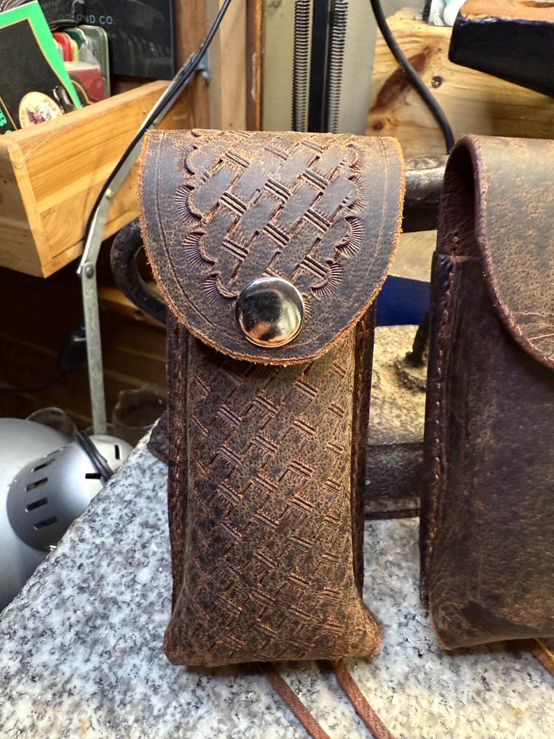K&ouml;nnte beinhalten: Eine braune Ledertasche mit einem gewebten Muster und einer dekorativen Klappe, die mit einem silberfarbenen Knopf befestigt ist. Die Tasche ist f&uuml;r einen kleinen Gegenstand konzipiert und hat einen Riemen. Das Leder hat ein abgenutztes Aussehen.