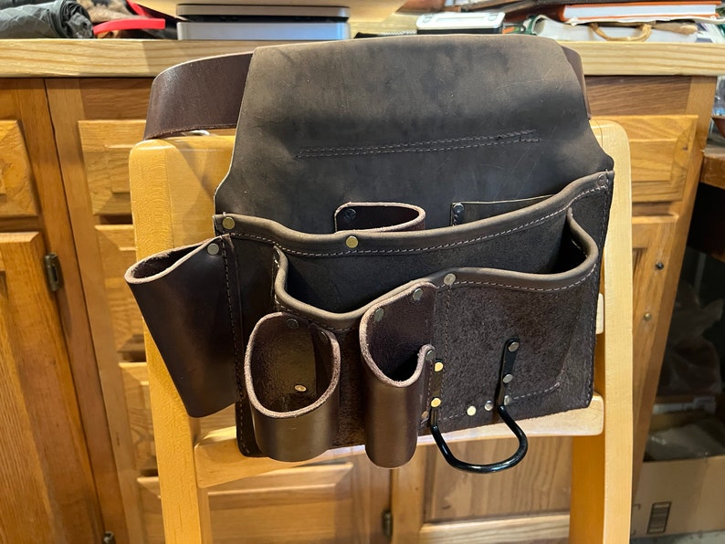 May include: A brown leather tool belt with multiple pockets and a black metal hook. The belt is hanging on a wooden chair.