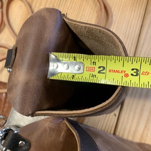 May include: A brown leather pouch with a yellow measuring tape inside. The tape reads "25ft" and "2 ft" with the Stanley logo visible. The pouch is open and the tape is measuring the inside of the pouch.