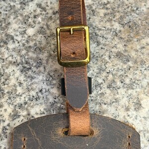 Op de afbeelding: Close-up van een bruine leren bagagelabel met een messing gesp en riem. Het label heeft een verweerde uitstraling met zichtbare stiksels en een getextureerd oppervlak. De riem is met een messing gesp aan het label bevestigd.