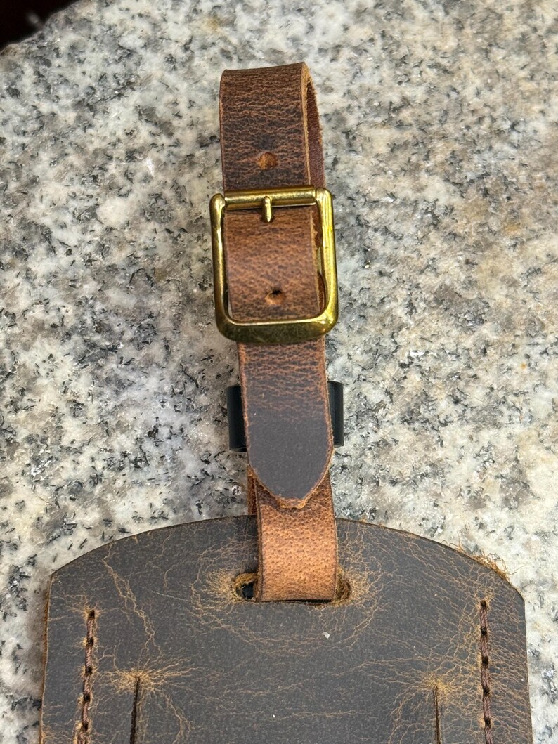 Op de afbeelding: Close-up van een bruine leren bagagelabel met een messing gesp en riem. Het label heeft een verweerde uitstraling met zichtbare stiksels en een getextureerd oppervlak. De riem is met een messing gesp aan het label bevestigd.