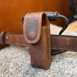 May include: A brown leather belt pouch with a flap closure, attached to a brown leather belt. The pouch has a distressed finish, and the belt is secured with a metal buckle. The pouch is designed to be worn on the waist.