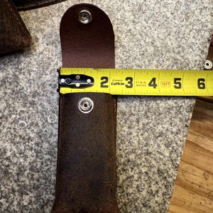 K&ouml;nnte beinhalten: Eine braune Lederscheide mit Druckknopfverschluss, etwa 12,7 cm lang. Die Scheide ist offen und gibt den Blick ins Innere frei. Ein gelbes Ma&szlig;band wird verwendet, um die L&auml;nge der Scheide zu zeigen. Das Leder hat ein Used-Aussehen.