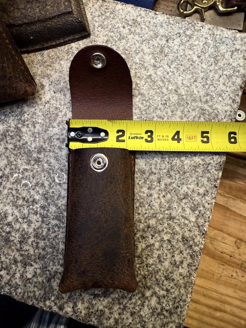 K&ouml;nnte beinhalten: Eine braune Lederscheide mit Druckknopfverschluss, etwa 12,7 cm lang. Die Scheide ist offen und gibt den Blick ins Innere frei. Ein gelbes Ma&szlig;band wird verwendet, um die L&auml;nge der Scheide zu zeigen. Das Leder hat ein Used-Aussehen.